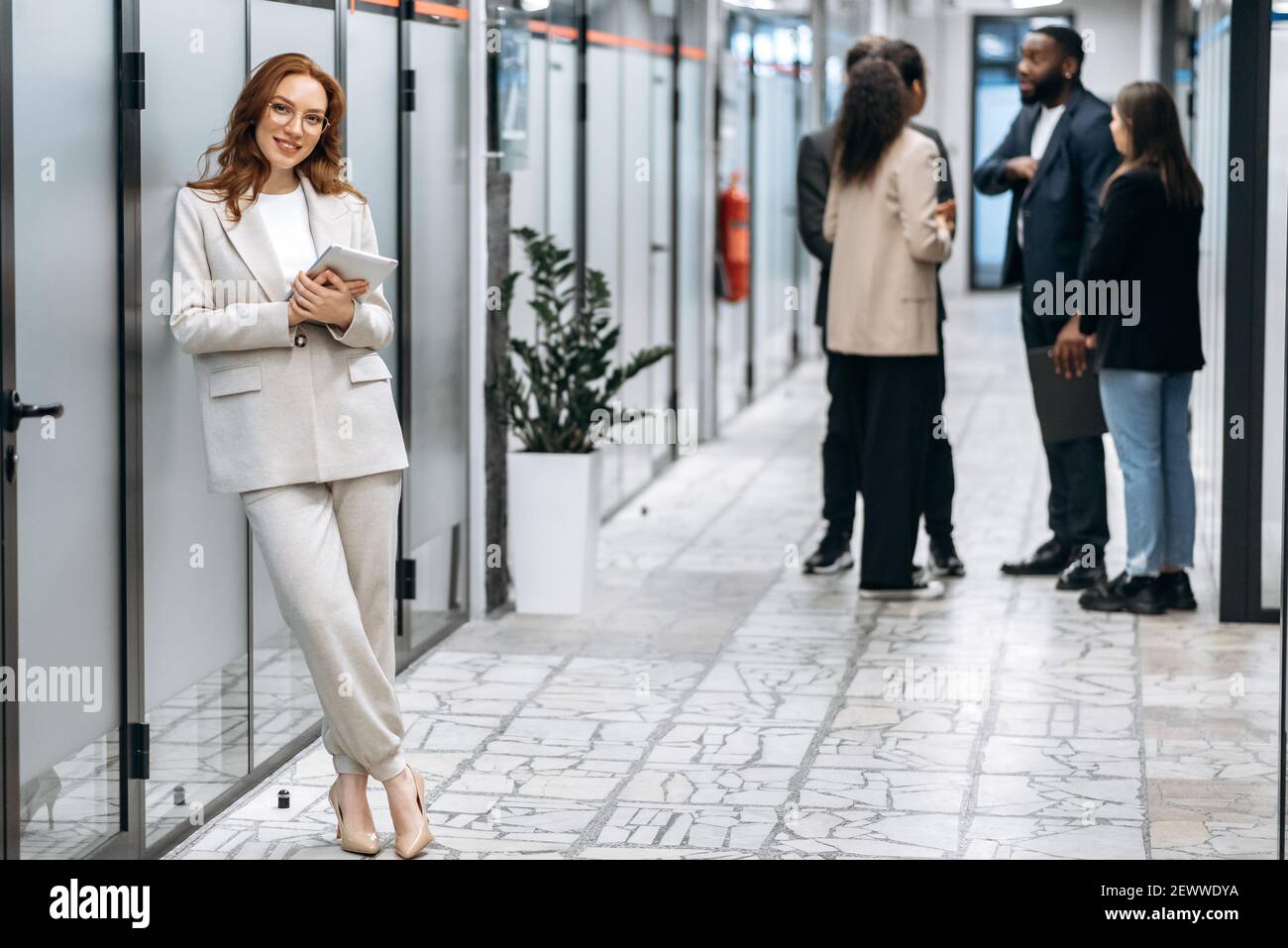 Splendida donna d'affari di successo in elegante abito elegante è in piedi nella sala d'ufficio, sorridente. Attraente dipendente rosso che guarda la fotocamera, tieni il tablet in braccio Foto Stock