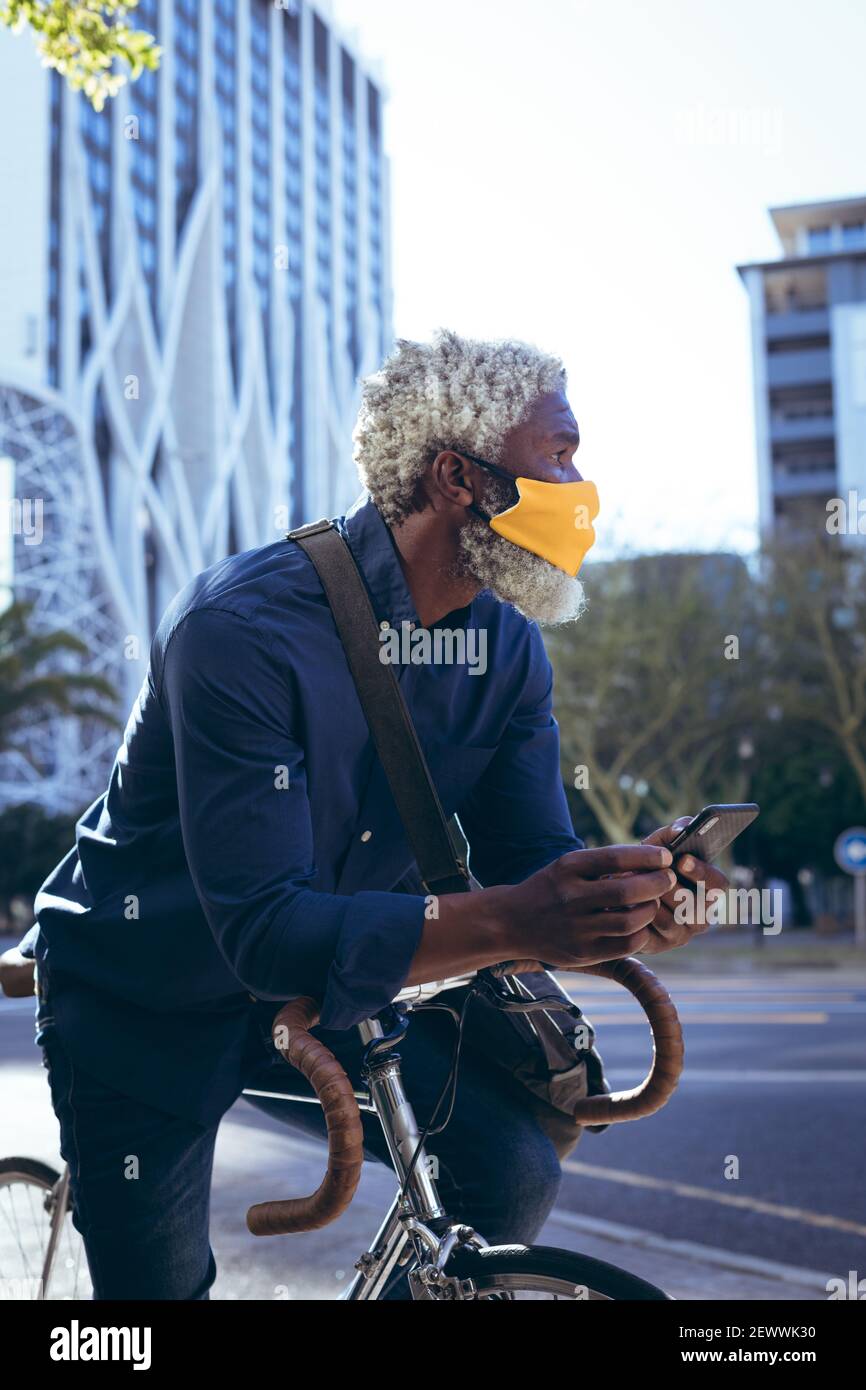 Uomo anziano afro-americano che indossa una maschera seduta in bicicletta in strada utilizzando lo smartphone Foto Stock