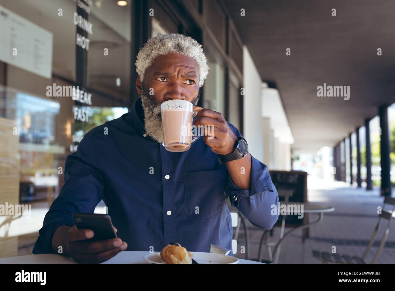 Uomo anziano afroamericano seduto al tavolo all'esterno del caffè che beve caffè utilizzando smartphone e looki Foto Stock