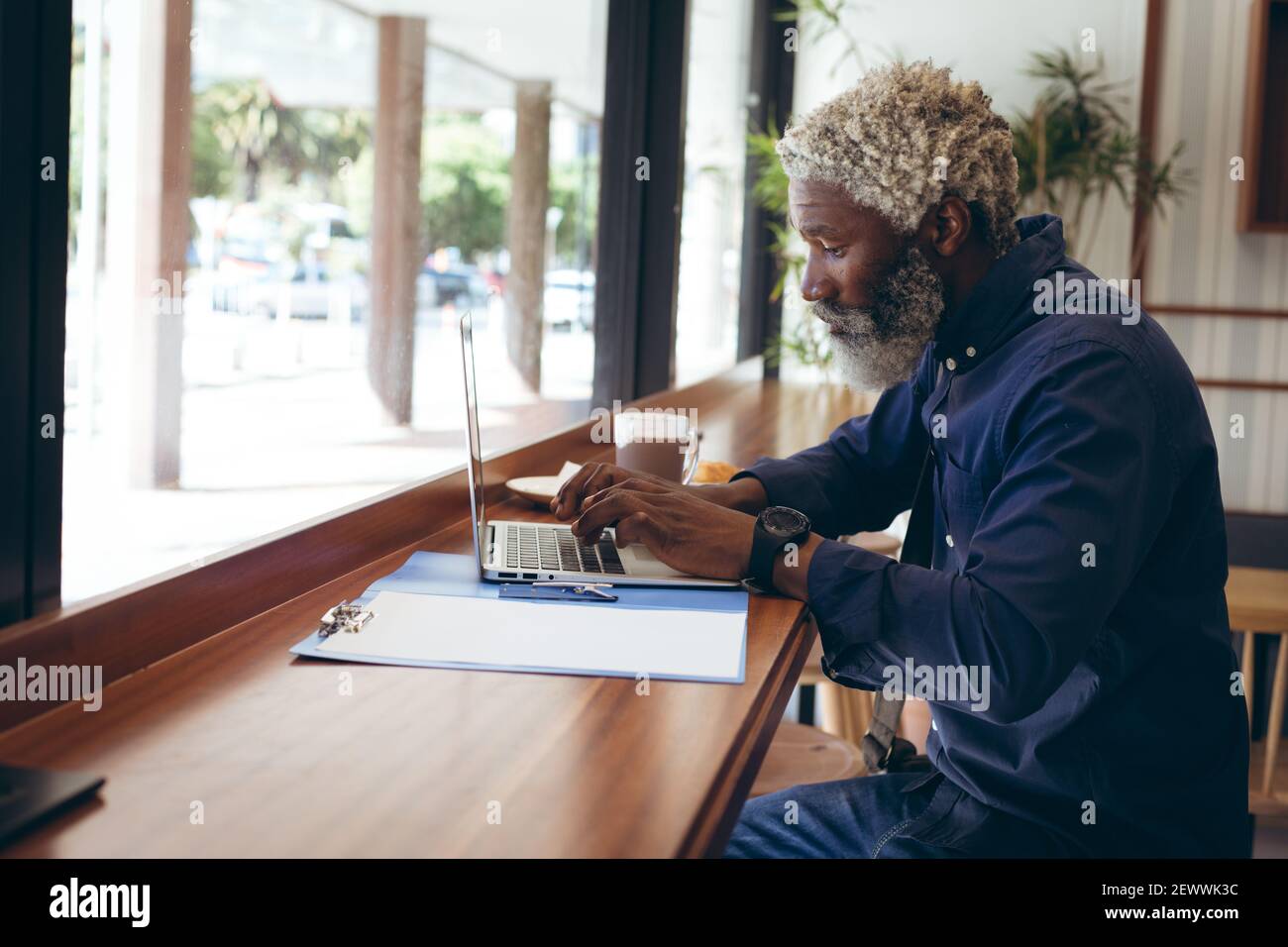 Uomo anziano afroamericano seduto al tavolo in un bar che lavora utilizzando un computer portatile Foto Stock