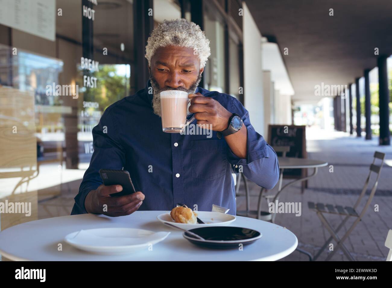 Uomo anziano afroamericano seduto al tavolo all'esterno del caffè che beve caffè e utilizzo dello smartphone Foto Stock