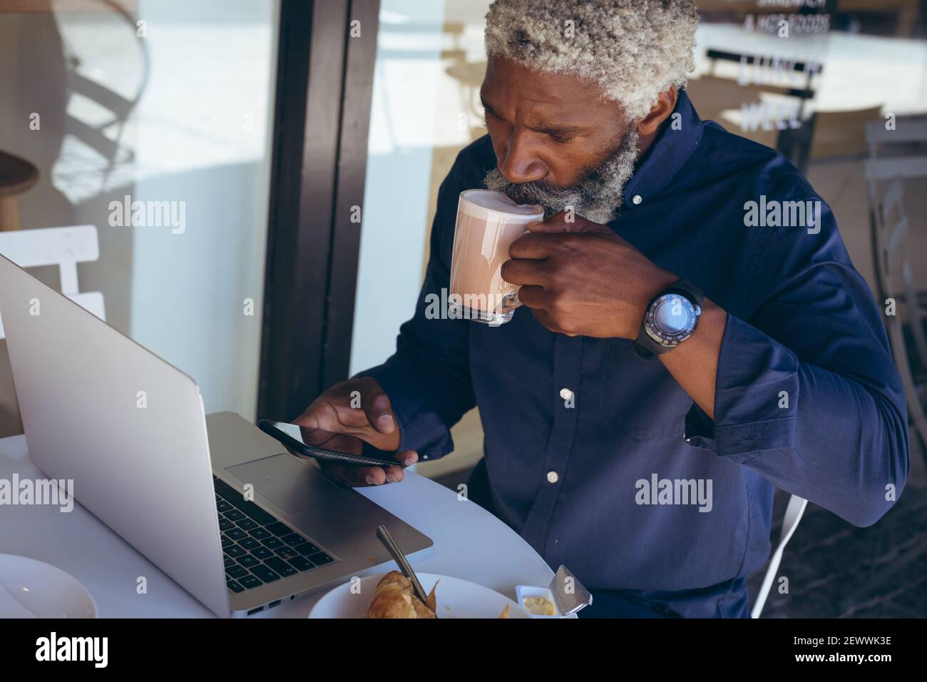 Uomo anziano afroamericano seduto al tavolo all'esterno del caffè che beve caffè con computer portatile e smartphon Foto Stock