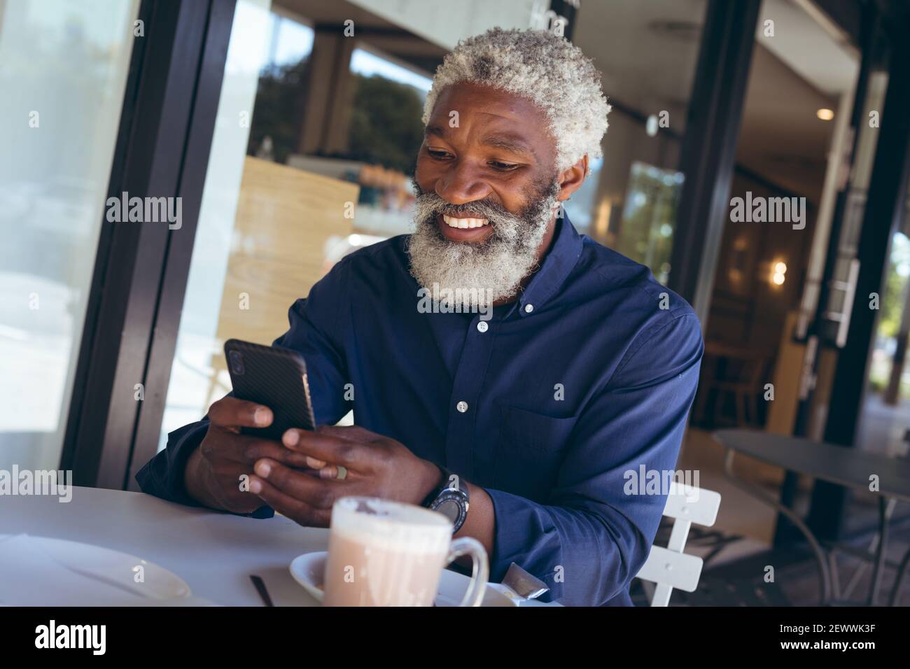 Uomo anziano afroamericano seduto al tavolo fuori del cafe con caffè utilizzando smartphone e sorridendo Foto Stock