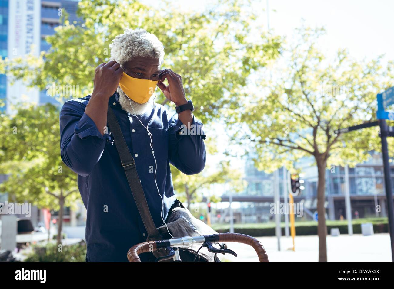 Uomo anziano afro-americano che indossa una maschera seduta in bicicletta in strada indossare gli auricolari Foto Stock