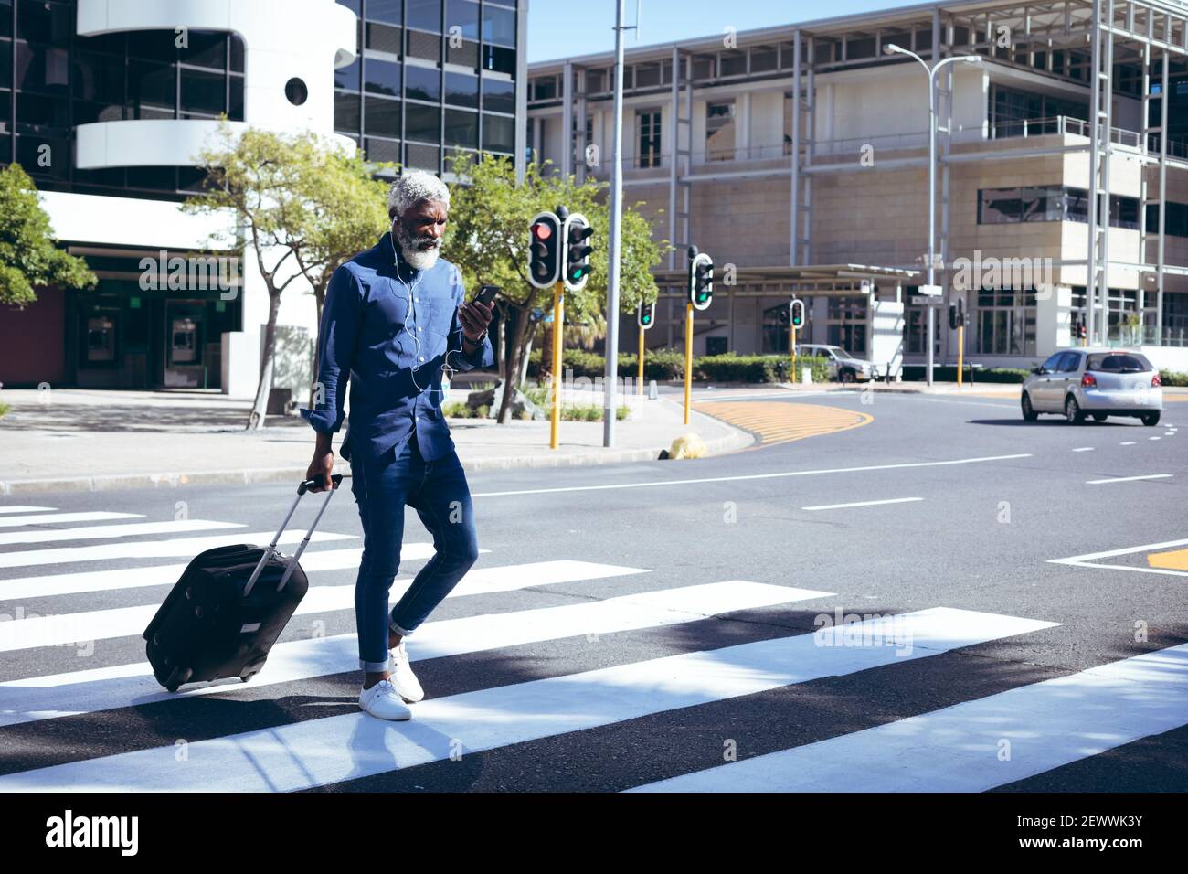 Uomo anziano afro-americano che indossa auricolari e utilizza la ruota per smartphone valigia dall'altra parte della strada Foto Stock