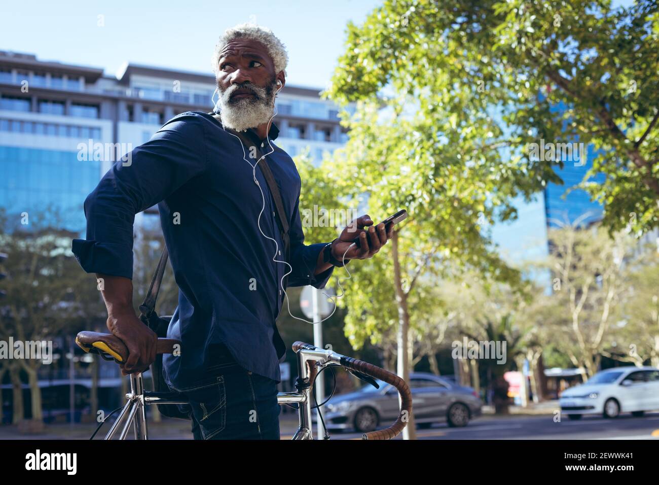 Uomo anziano afro-americano che indossa auricolari seduti in bicicletta via utilizzando lo smartphone Foto Stock