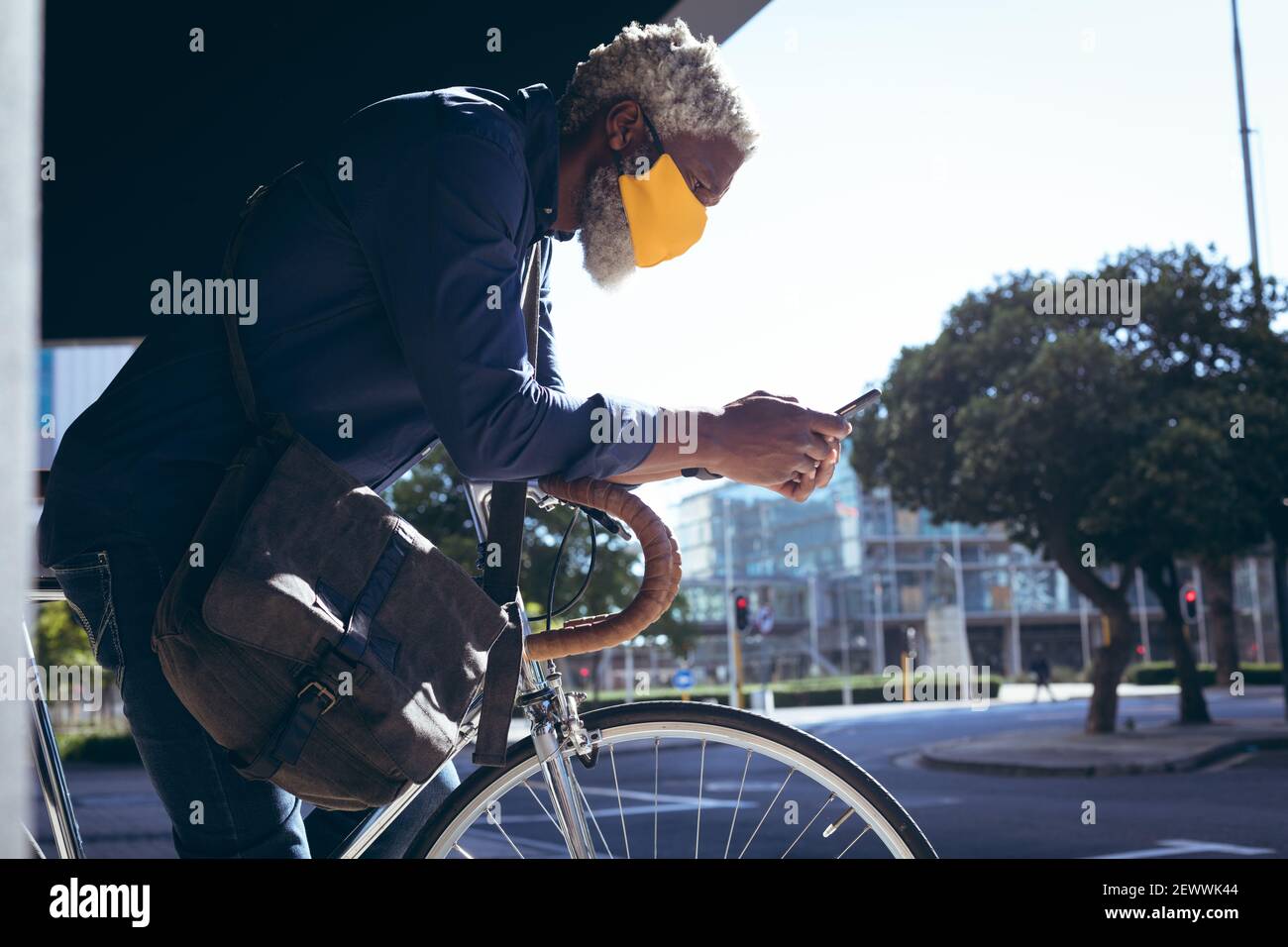 Uomo anziano afro-americano che indossa la maschera che pende sulla bicicletta in strada utilizzando lo smartphone Foto Stock