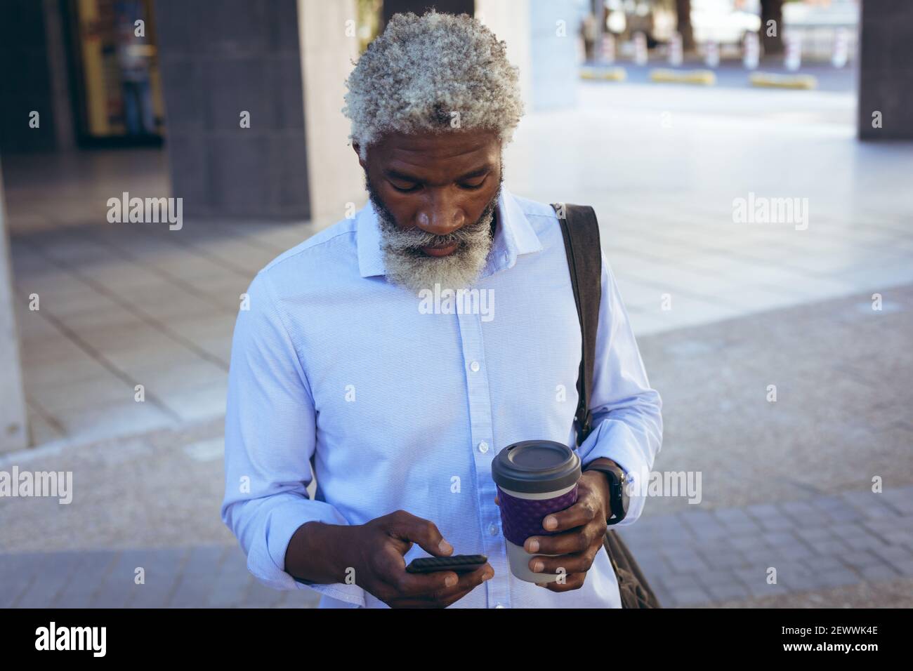 L'uomo anziano afroamericano che sta in piedi nella strada che tiene il caffè e. tramite smartphone Foto Stock