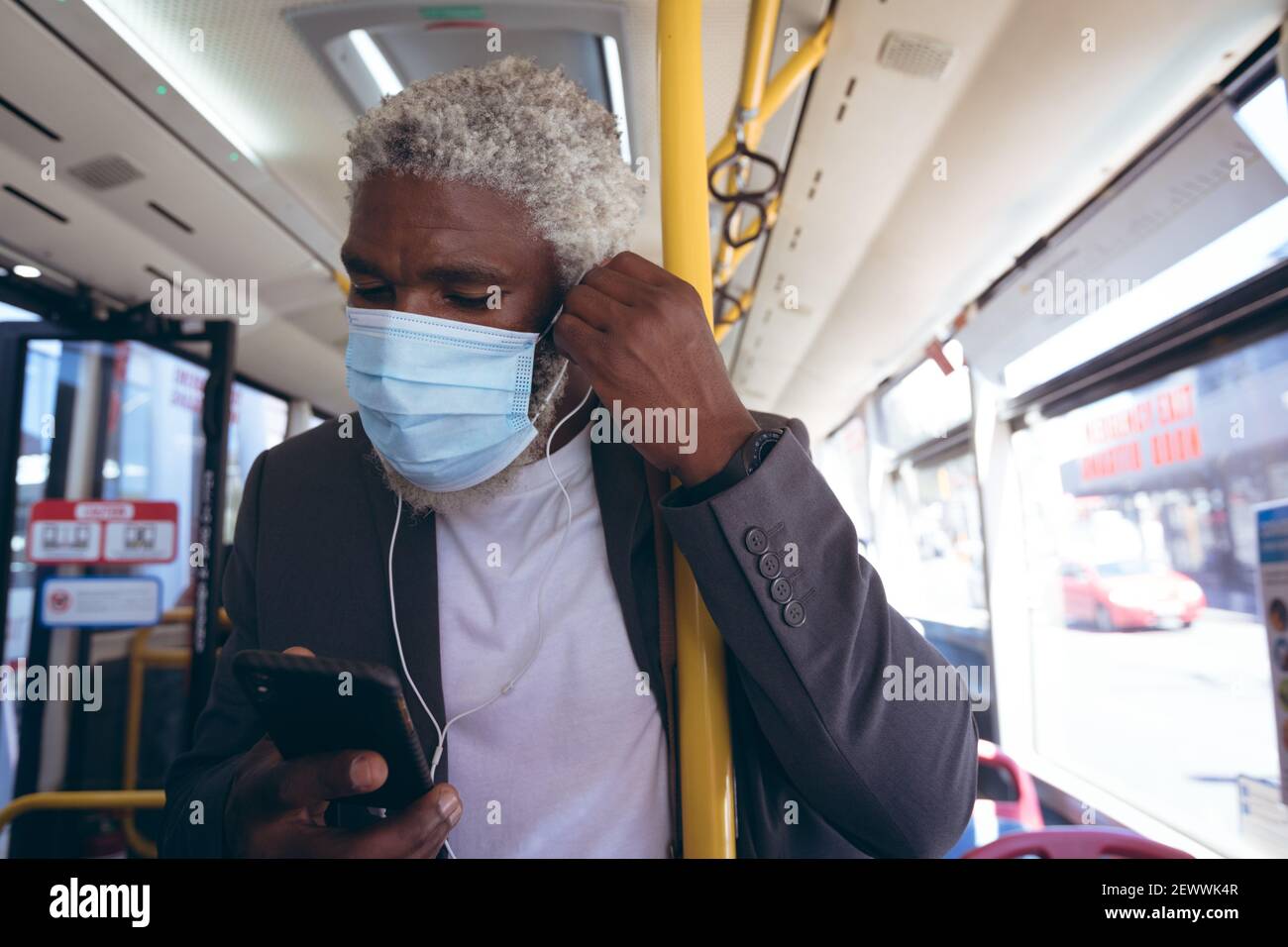 Uomo anziano afro-americano che indossa una maschera per il viso indossando gli auricolari stare in autobus utilizzando lo smartphone Foto Stock