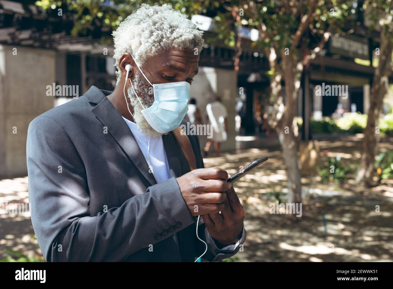 Uomo anziano afroamericano che indossa maschera facciale e auricolari in piedi in strada utilizzando lo smartphone Foto Stock