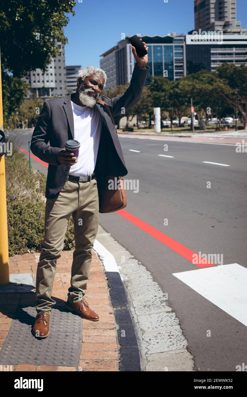 Uomo anziano afroamericano in piedi a bordo strada che tiene il caffè e. smartphone che salutano un taxi Foto Stock