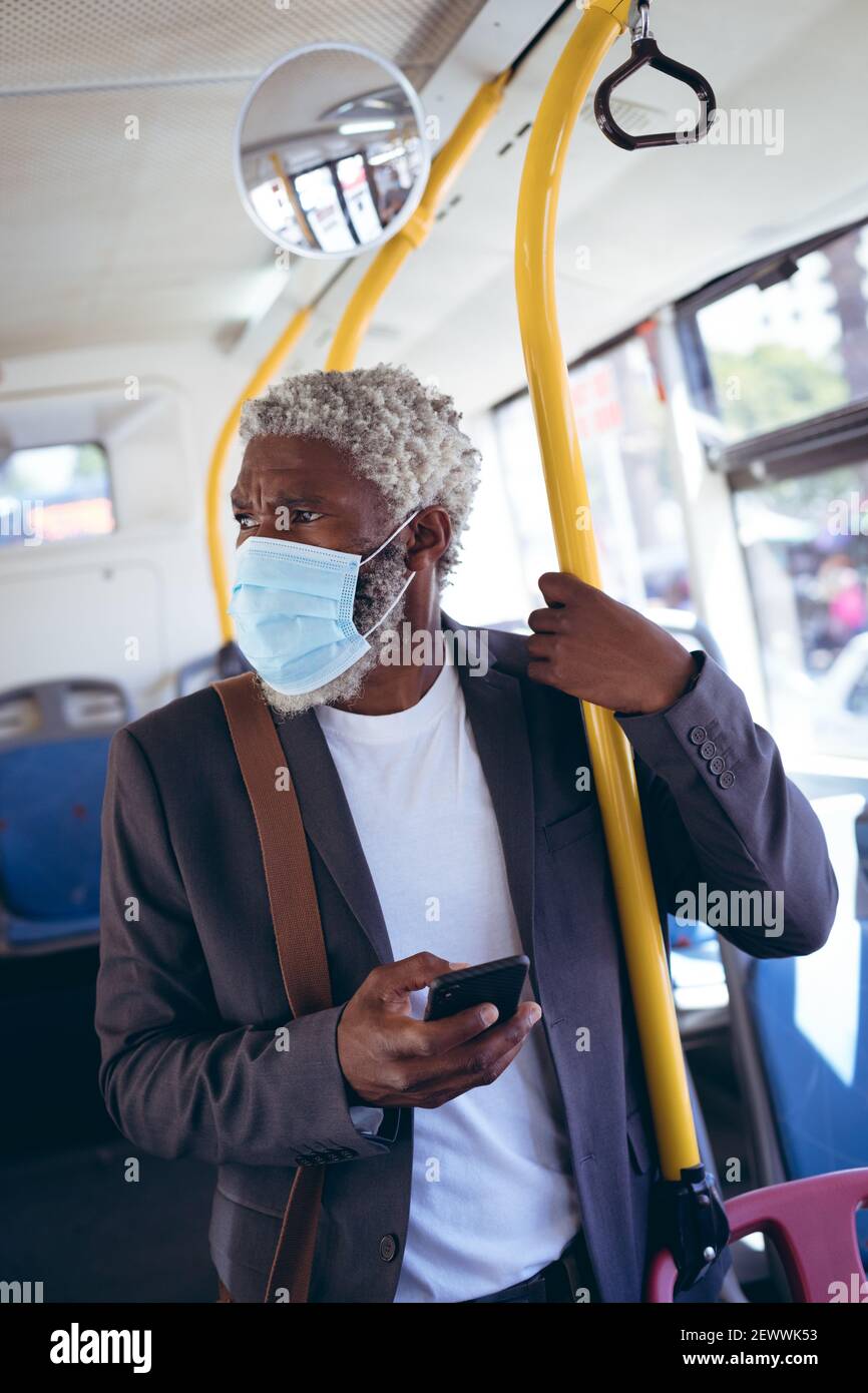 Uomo anziano afro-americano che indossa una maschera facciale in piedi sull'autobus tenere lo smartphone Foto Stock