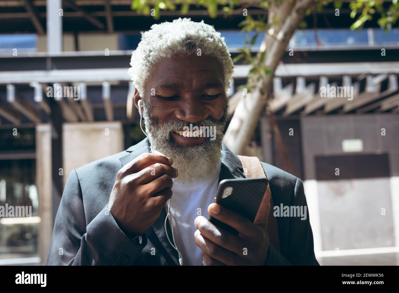 Uomo anziano afroamericano che indossa auricolari in piedi in strada utilizzando smartphone e sorridente Foto Stock