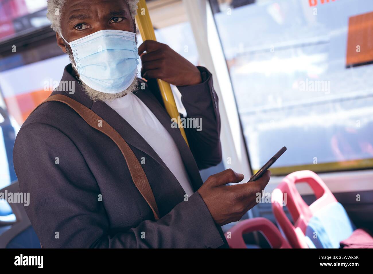 Uomo anziano afro-americano che indossa una maschera facciale in piedi sull'autobus tramite smartphone Foto Stock