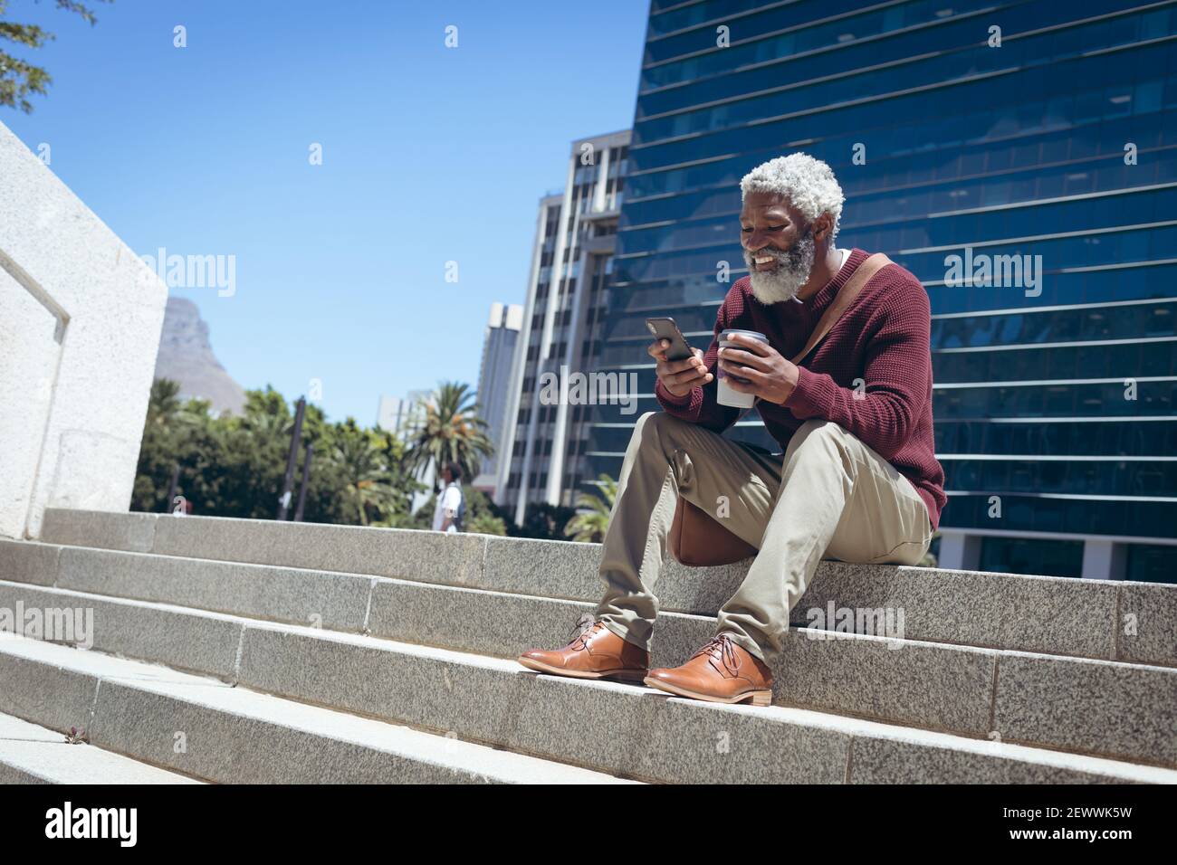 Uomo anziano afroamericano seduto sui gradini fuori della tenuta dell'edificio caffè con smartphone e sm Foto Stock