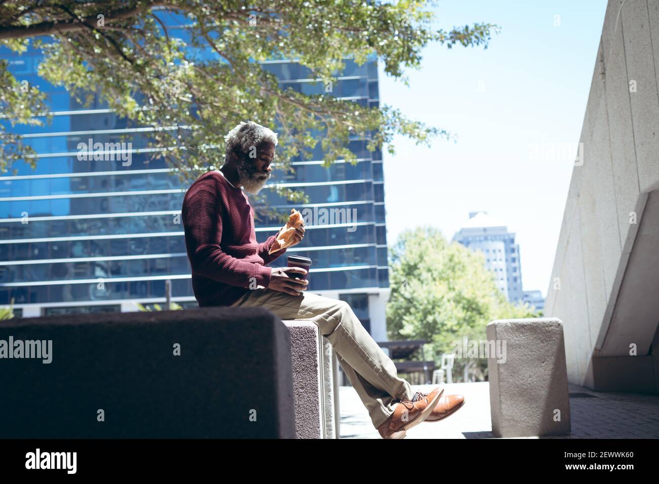 Uomo anziano afro-americano seduto sul muro in strada mangiare panino e caffè Foto Stock