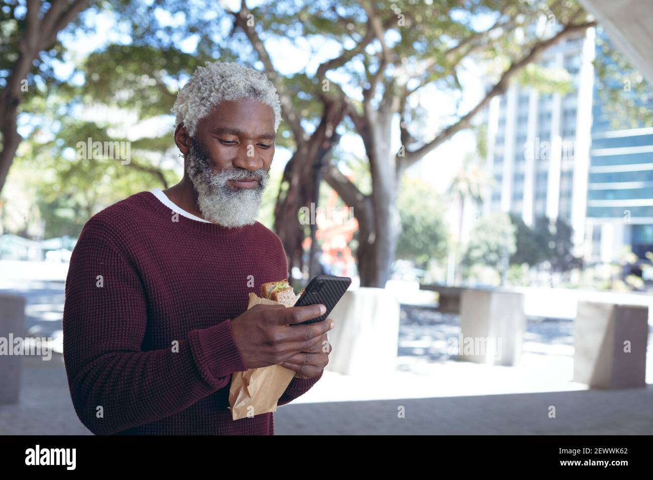 Uomo anziano afroamericano in strada mangiando sandwich e usando smartphone Foto Stock