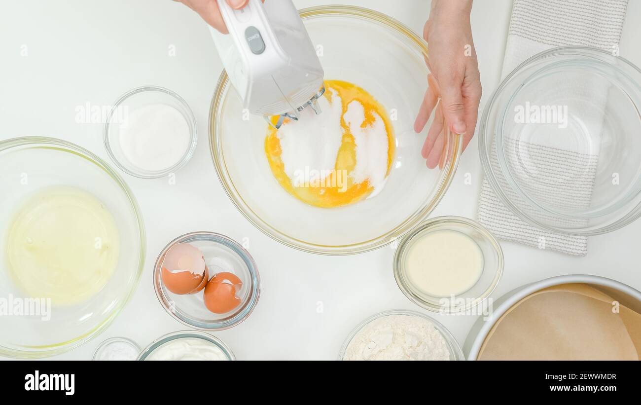 Le mani della donna mescolano i tuorli d'uovo con lo zucchero in una ciotola di vetro usando un miscelatore elettrico. Primo piano cottura, piatto Foto Stock