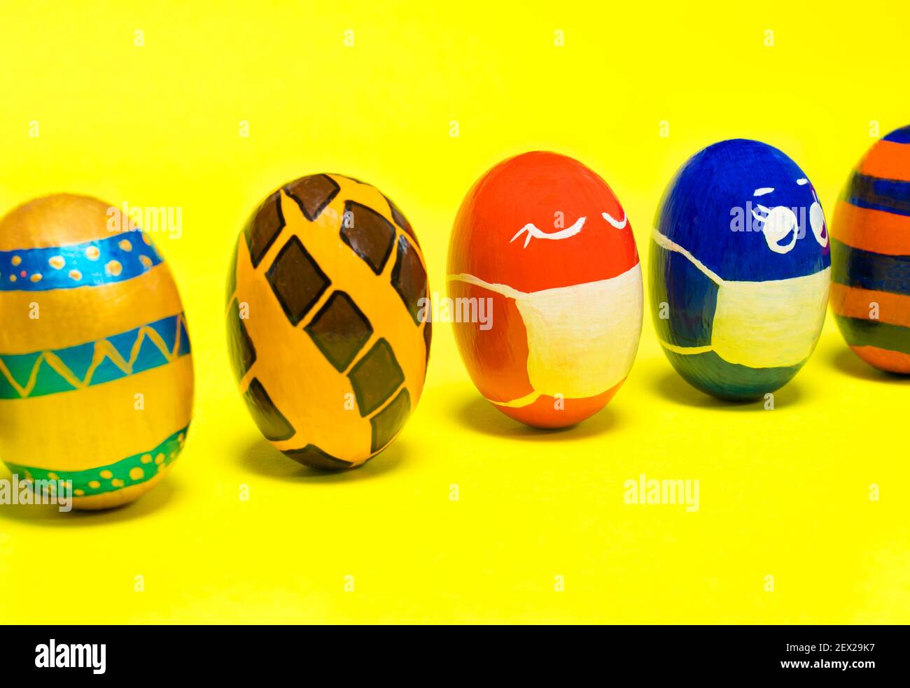 Uova di Pasqua multicolore su sfondo giallo. Uova con volti messi su maschere protettive contro il coronavirus durante la quarantena. Foto Stock