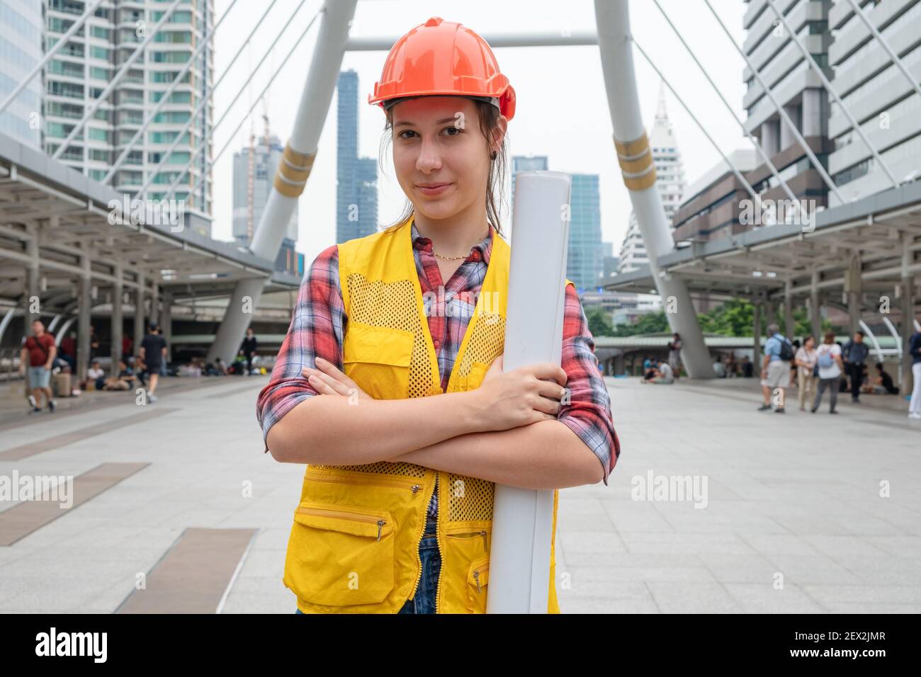 Donna caucasica costruttore in giubbotto di sicurezza con casco a braccia incrociate e la disposizione di cantiere di tenuta nell'urbano Foto Stock