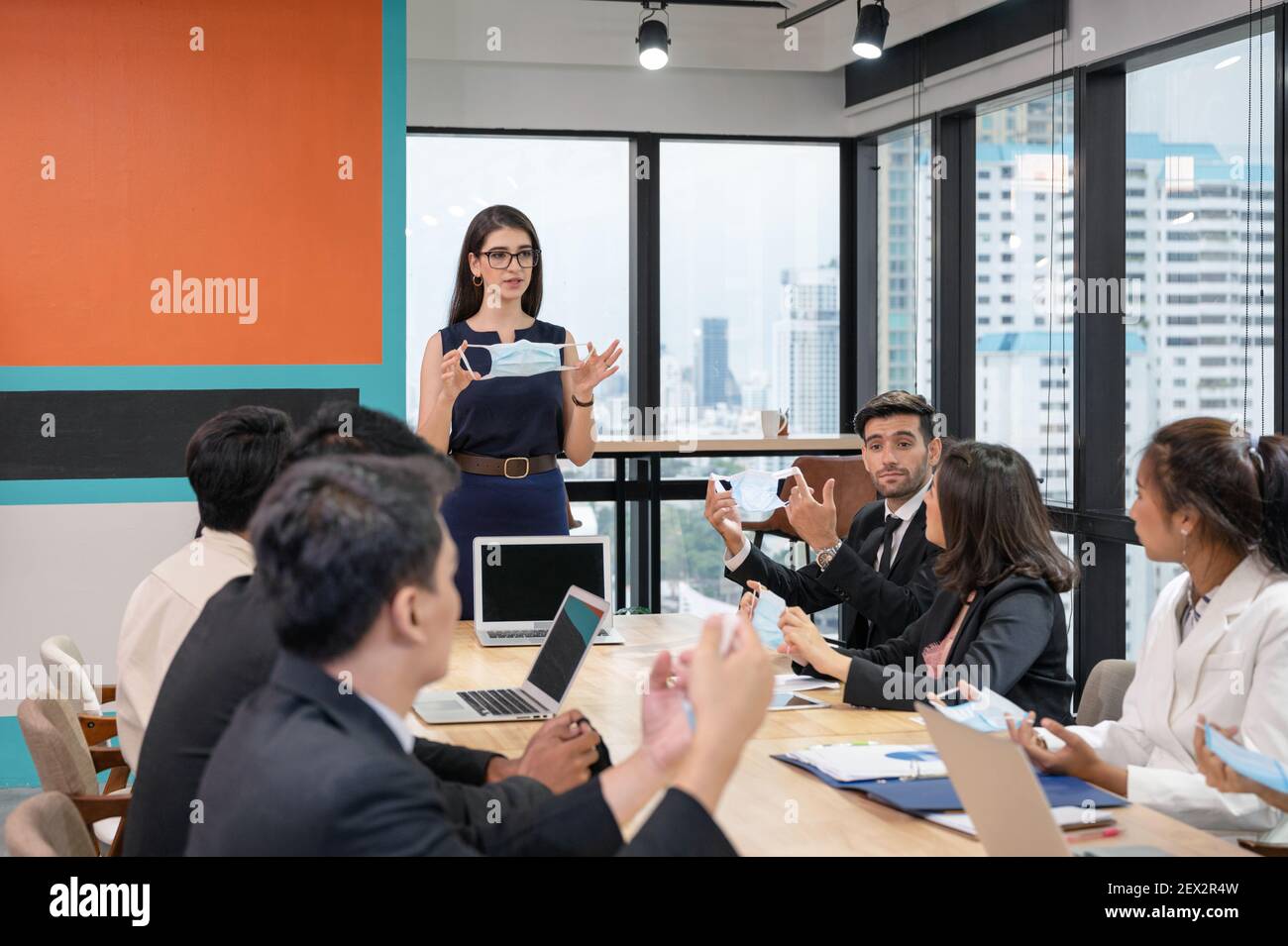 Donna dirigente caucasica che tiene maschera viso con presentare una nuova politica aziendale in un nuovo ufficio normale durante una riunione multietnica Foto Stock