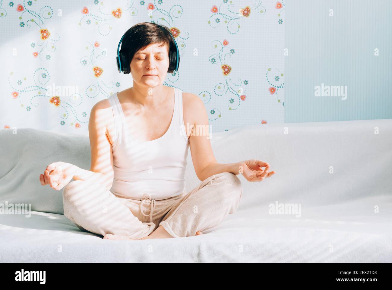 Una donna di mezza età si siede su un divano in posizione lotus con cuffie che ascoltano musica o lezioni di pratica spirituale su un computer portatile. Pratica di yoga Foto Stock