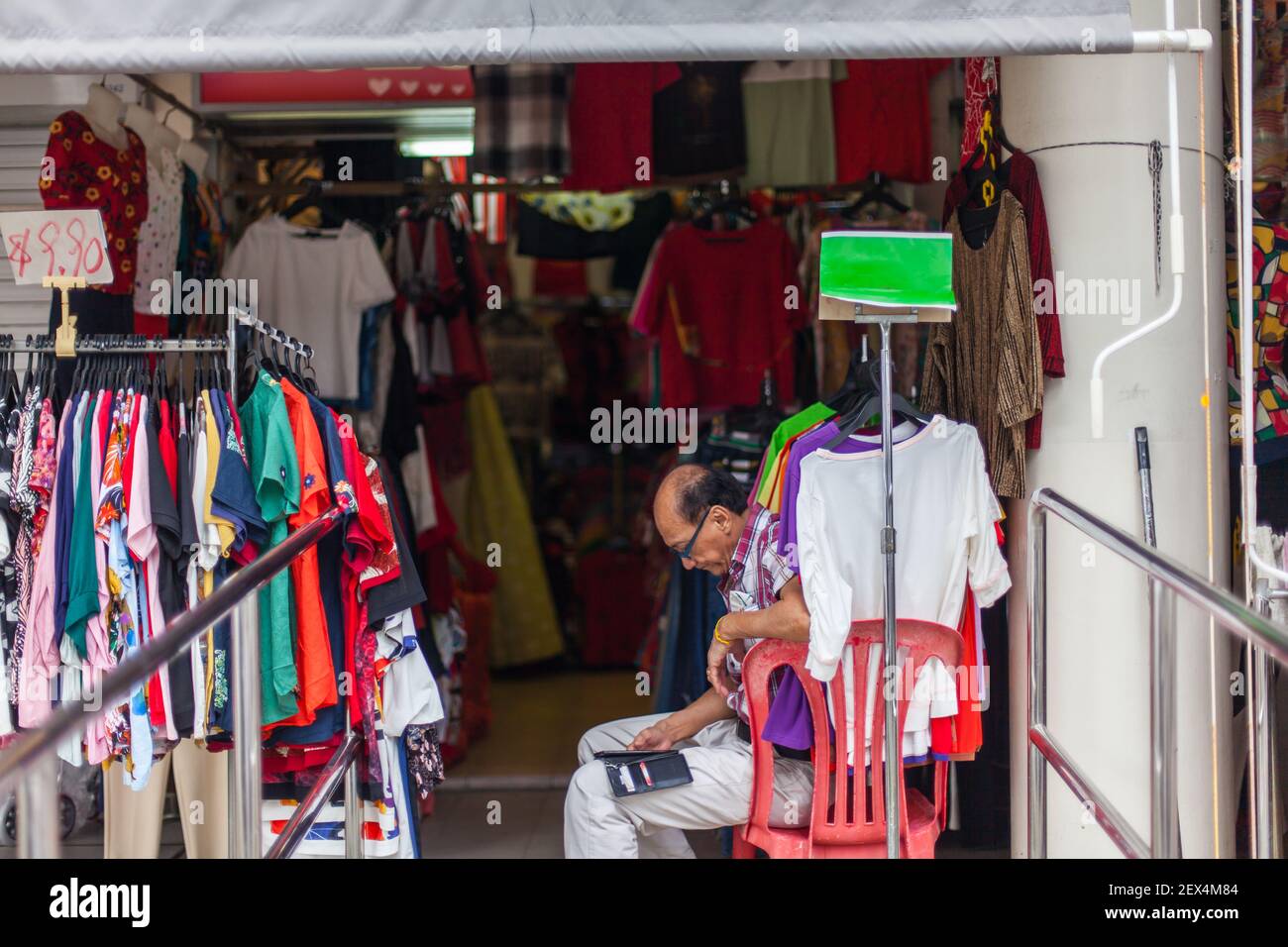 Un imprenditore cinese si siede al di fuori del negozio di abbigliamento di moda della sua donna indipendente, sta guardando sul suo telefono cellulare durante l'attesa. Singapore. Foto Stock