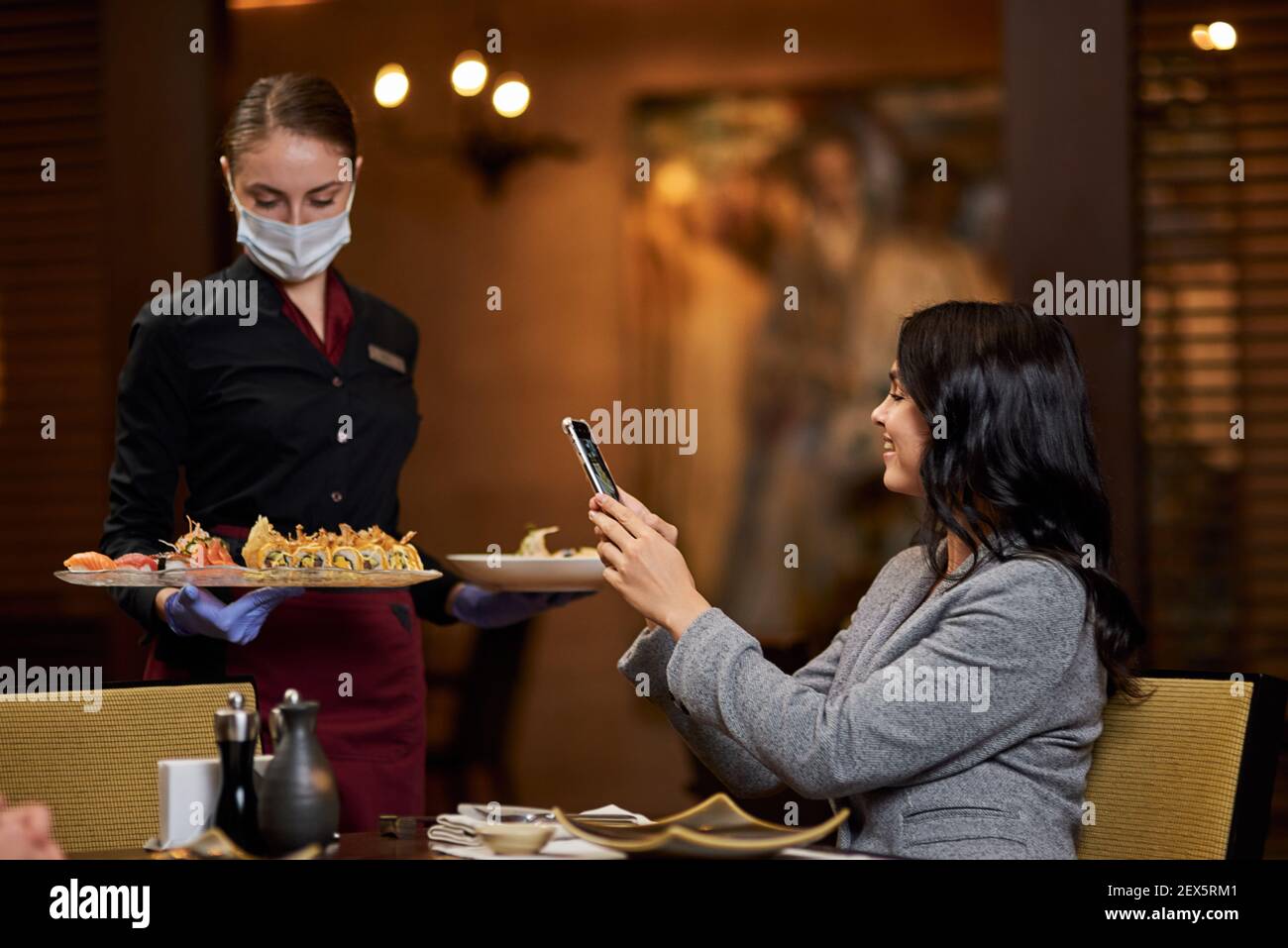 Cameriera seria in maschera protettiva del viso che consegna cibo ordine mentre femmina che ne fa una foto sul suo smartphone Foto Stock