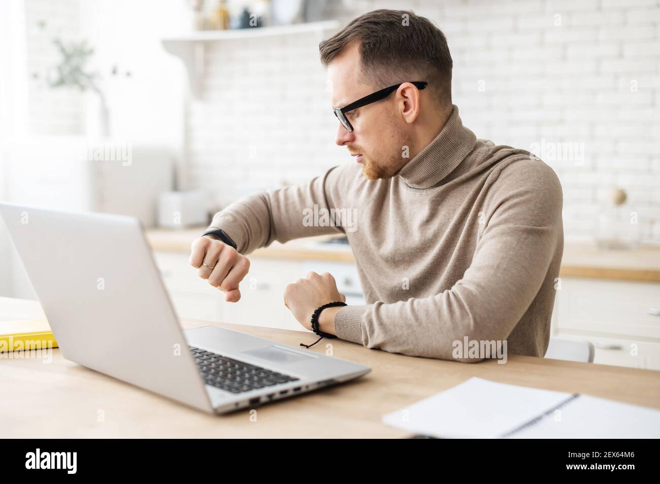 Un giovane imprenditore o insegnante affollato in occhiali seduti a casa e lavorando su un computer portatile, controllando il tempo da un orologio sul polso, il tempo per iniziare una conferenza online in streaming o una lezione virtuale Foto Stock