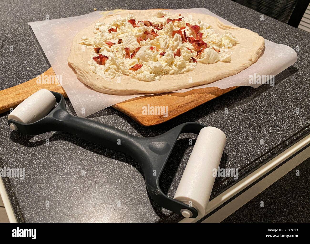 Pizza bianca fatta in casa con pancetta pronta per essere cucinata su una tavola di pizza accanto al rullo di impasto su un banco da cucina nero. Foto Stock