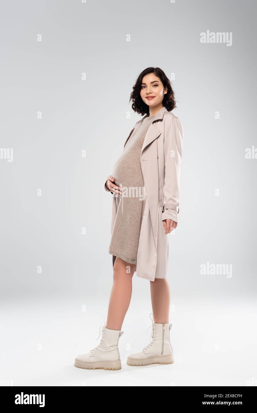 Donna incinta di stile con cappotto e stivali sorridenti sul grigio sfondo Foto Stock