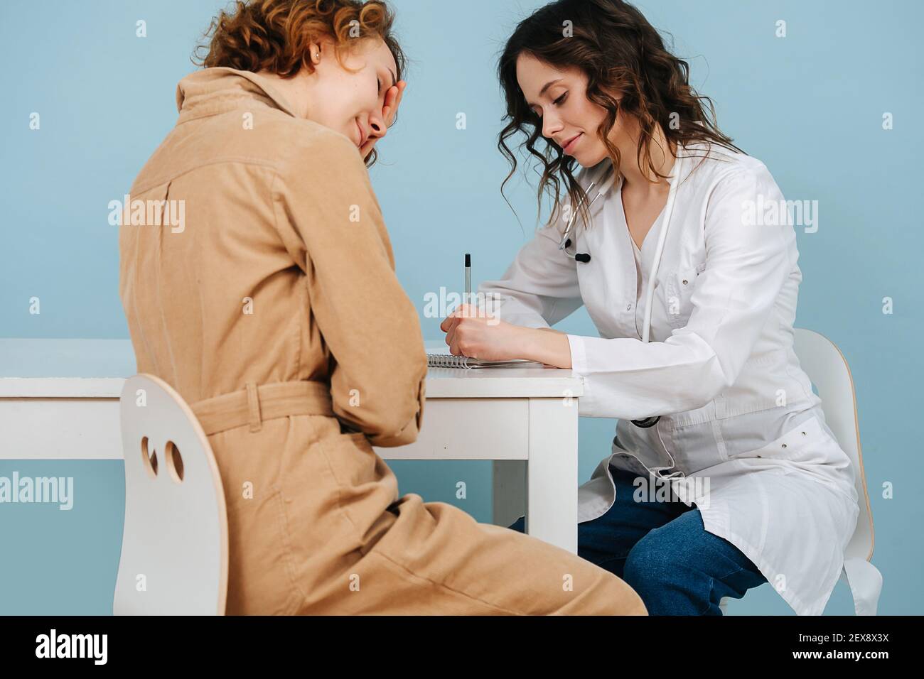 Donna in un ufficio medico, affetta da mal di testa, in attesa di un lavoro di documentazione. Su sfondo blu. Lei indossa veste bianca, jeans e stetoscopio. Foto Stock