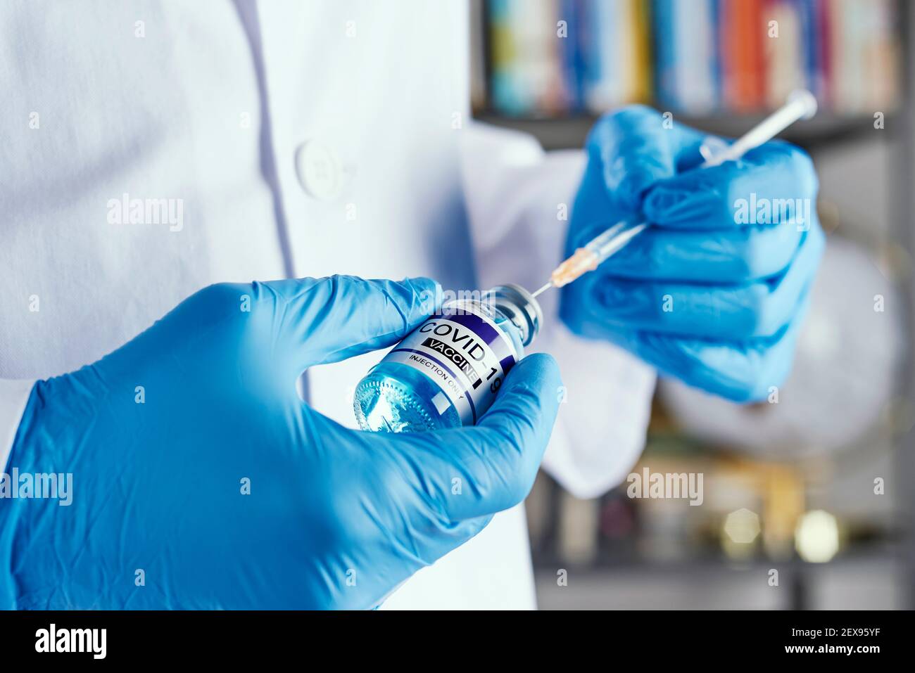 closeup di un operatore sanitario, indossando camice bianco e guanti chirurgici blu, riempire una siringa da un flaconcino simulato di vaccino covid-19, in un hou Foto Stock