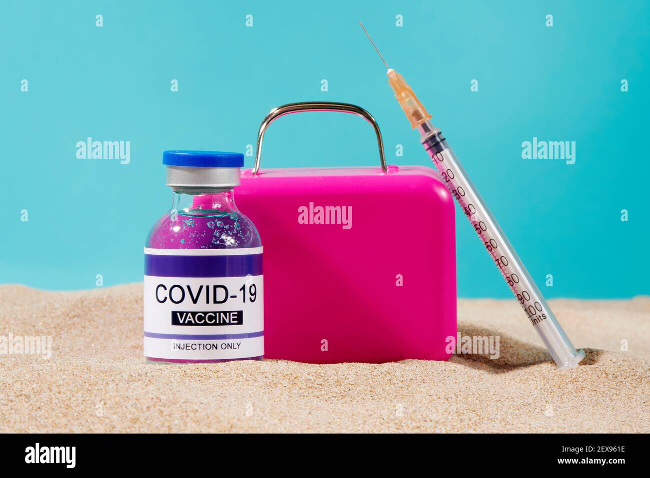 closeup di un flaconcino simulato di vaccino covid-19, una siringa e una valigia rosa sulla sabbia, su uno sfondo blu, raffigurante il turismo medico o tra Foto Stock