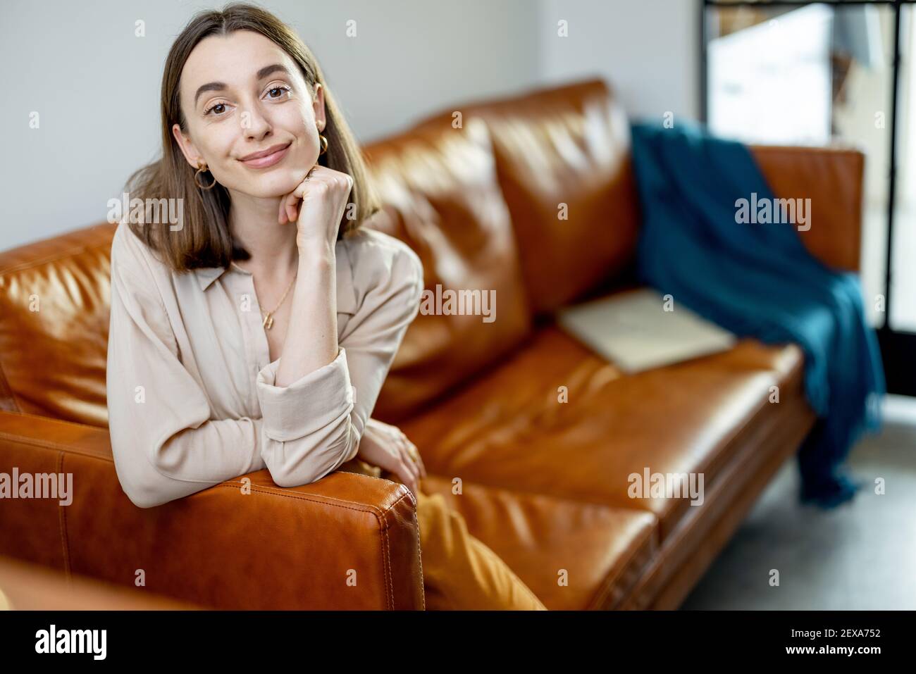 Ritratto di bella donna sorridente e guardando la macchina fotografica mentre si siede sul divano marrone confortevole su sfondo casa interno. Stile di vita e concetto di intimità domestica. Foto Stock