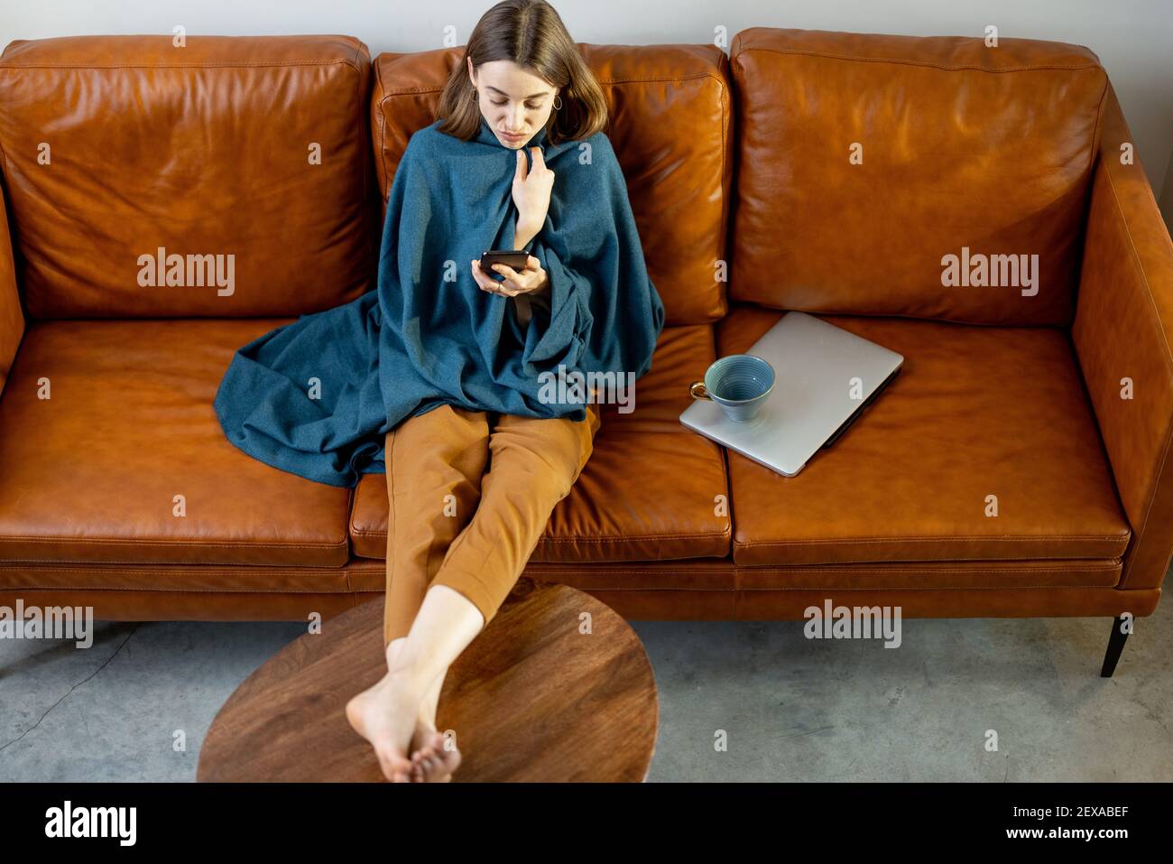 Bella donna con smartphone coperto da una plaid blu seduta sul divano marrone a casa. Stile di vita e concetto di intimità domestica. Foto Stock