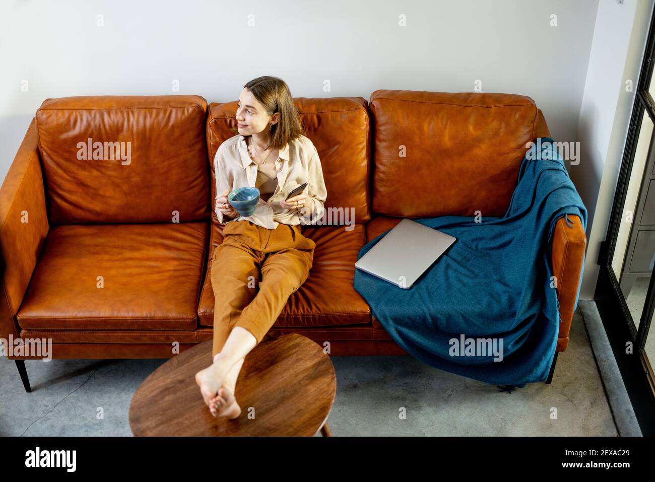 Bella donna con smartphone e tazza seduta sul divano marrone a casa. Stile di vita e concetto digitale. Foto Stock