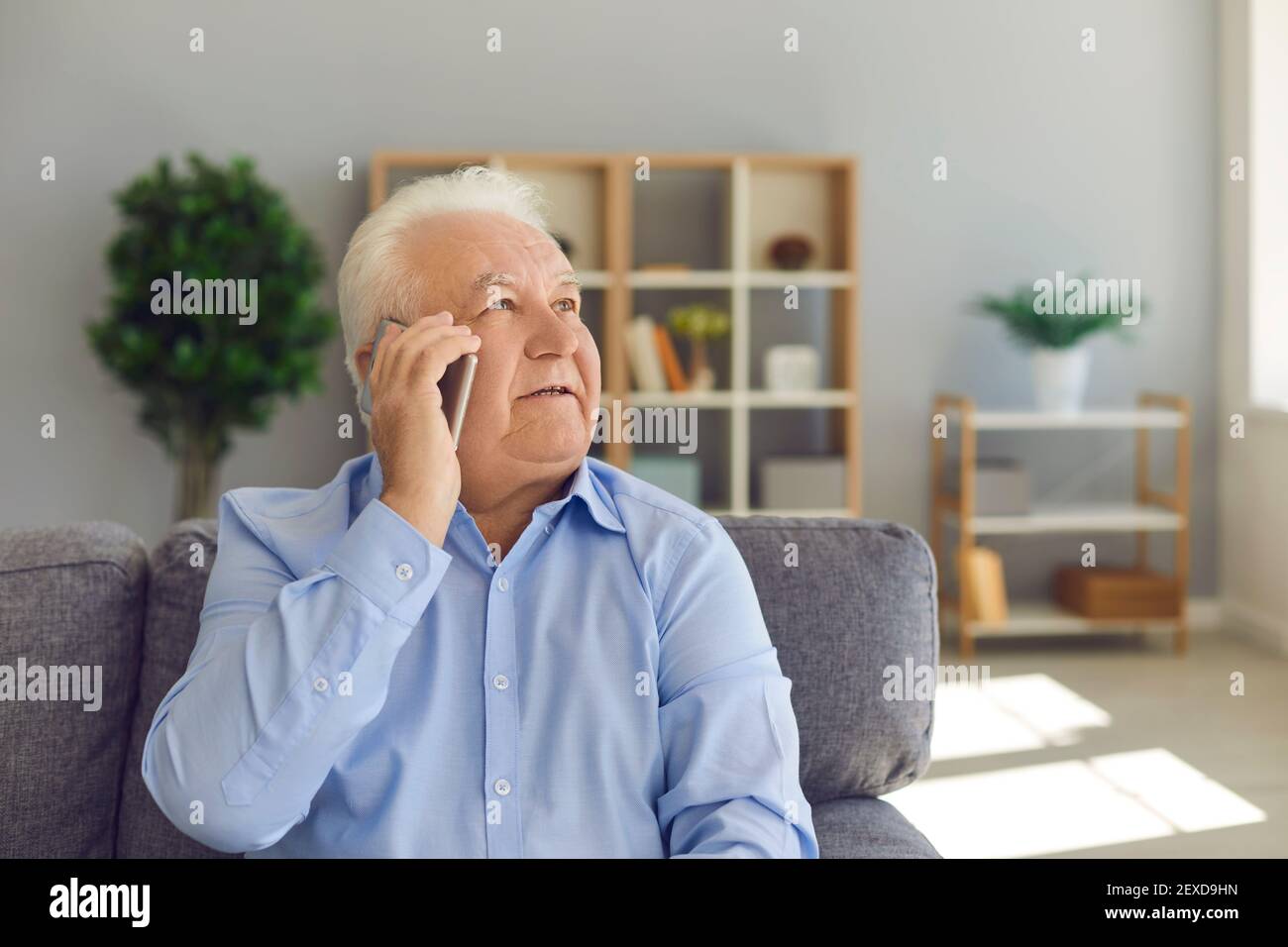 L'uomo anziano si siede su un divano in un soggiorno luminoso e parla al telefono con un attento sguardo in su. Foto Stock