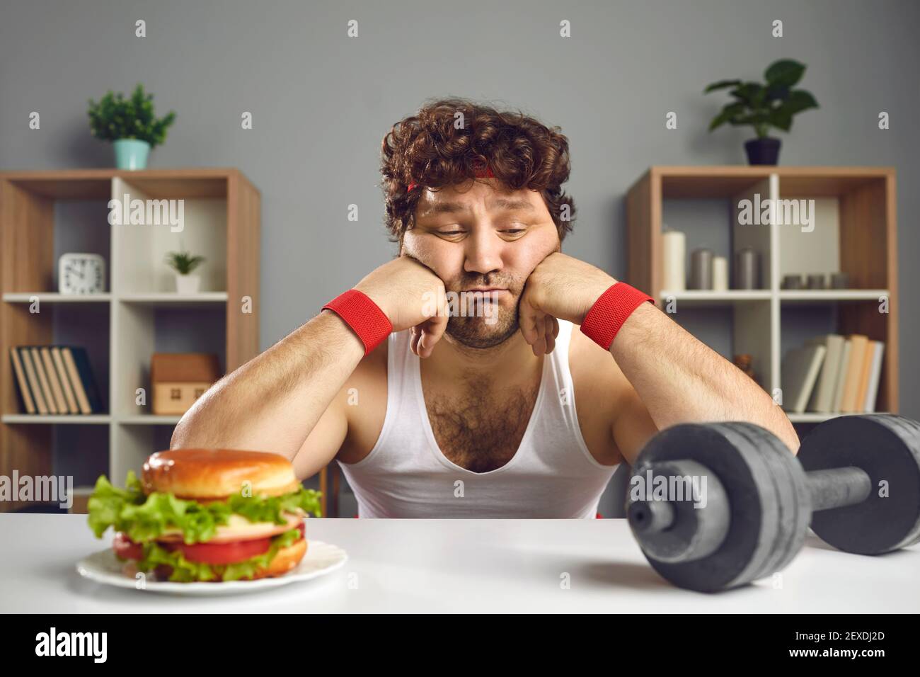 Uomo triste che guarda l'hamburger e dumbbell tentazione di scegliere tra cibo e formazione Foto Stock