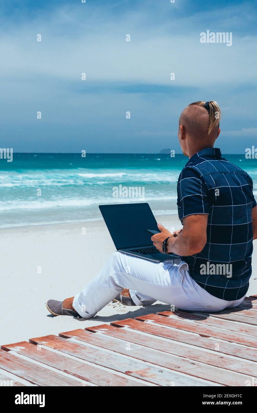 Giovane ragazzo di successo che guarda il mare, lavora con un computer portatile, seduto in riva al mare su una spiaggia di sabbia. Lavoro di uomo d'affari su un viaggio. Blogger, fre Foto Stock