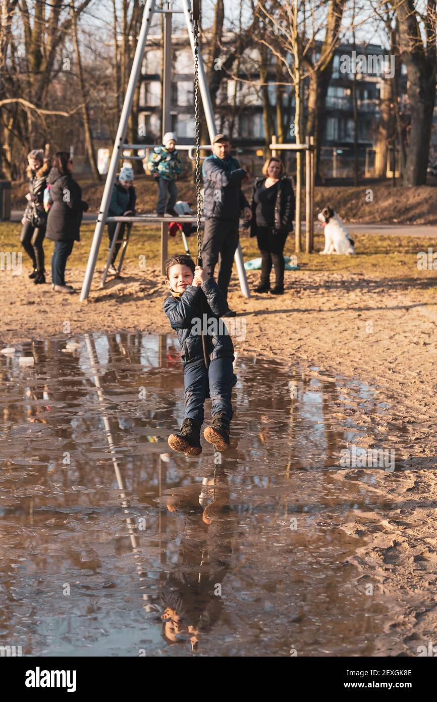 POZ, POLONIA - 21 febbraio 2021: Giovane ragazzo caucasico polacco felice che tiene su una zipline attrezzatura in un parco giochi nel Solacki parco con grande acqua puddle Foto Stock