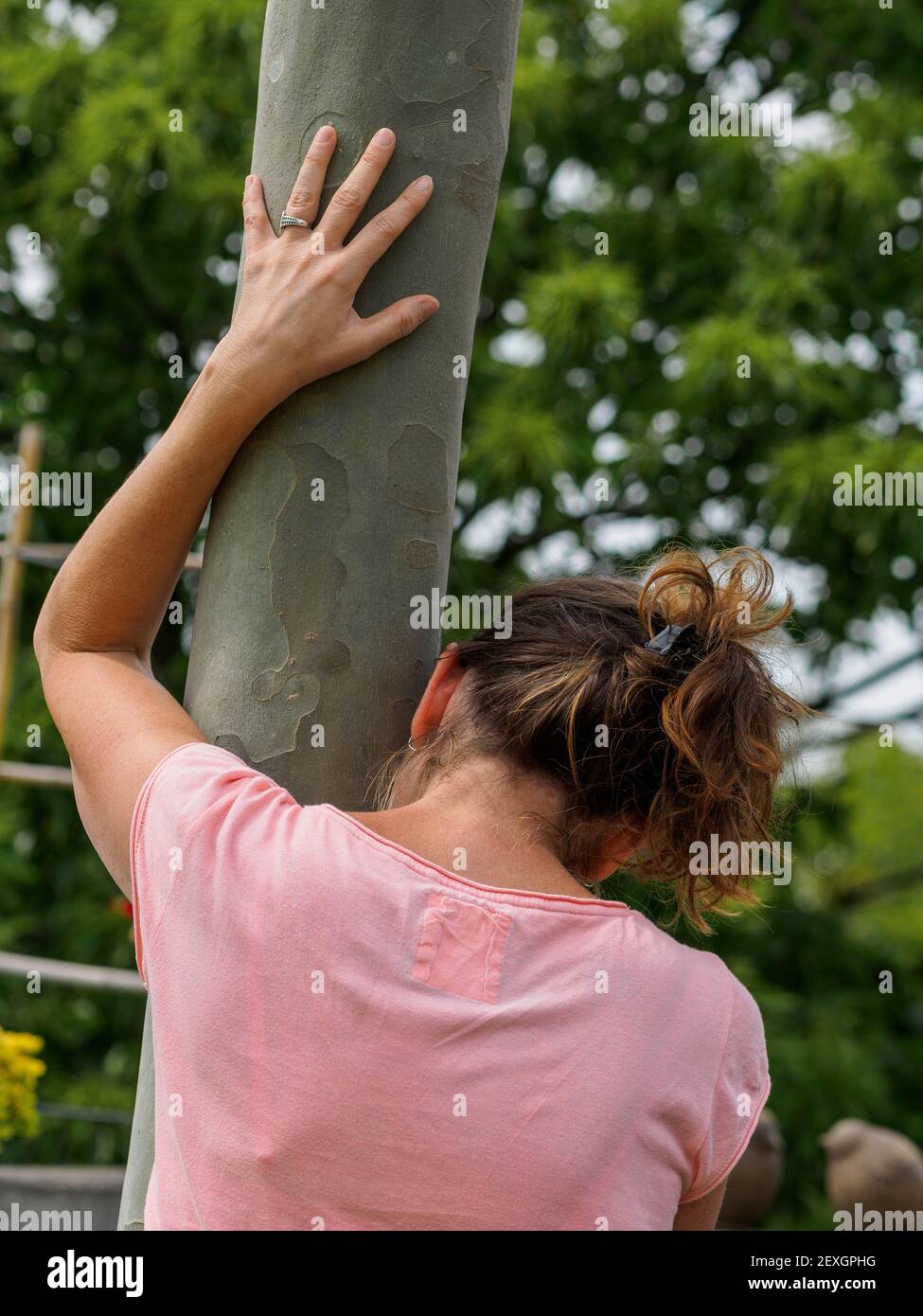 Giovane donna caucasica stanca si appoggia al tronco d'albero nel parco estivo. Tempo per rilassarsi. Concetto di salute mentale. Da dietro. Foto Stock