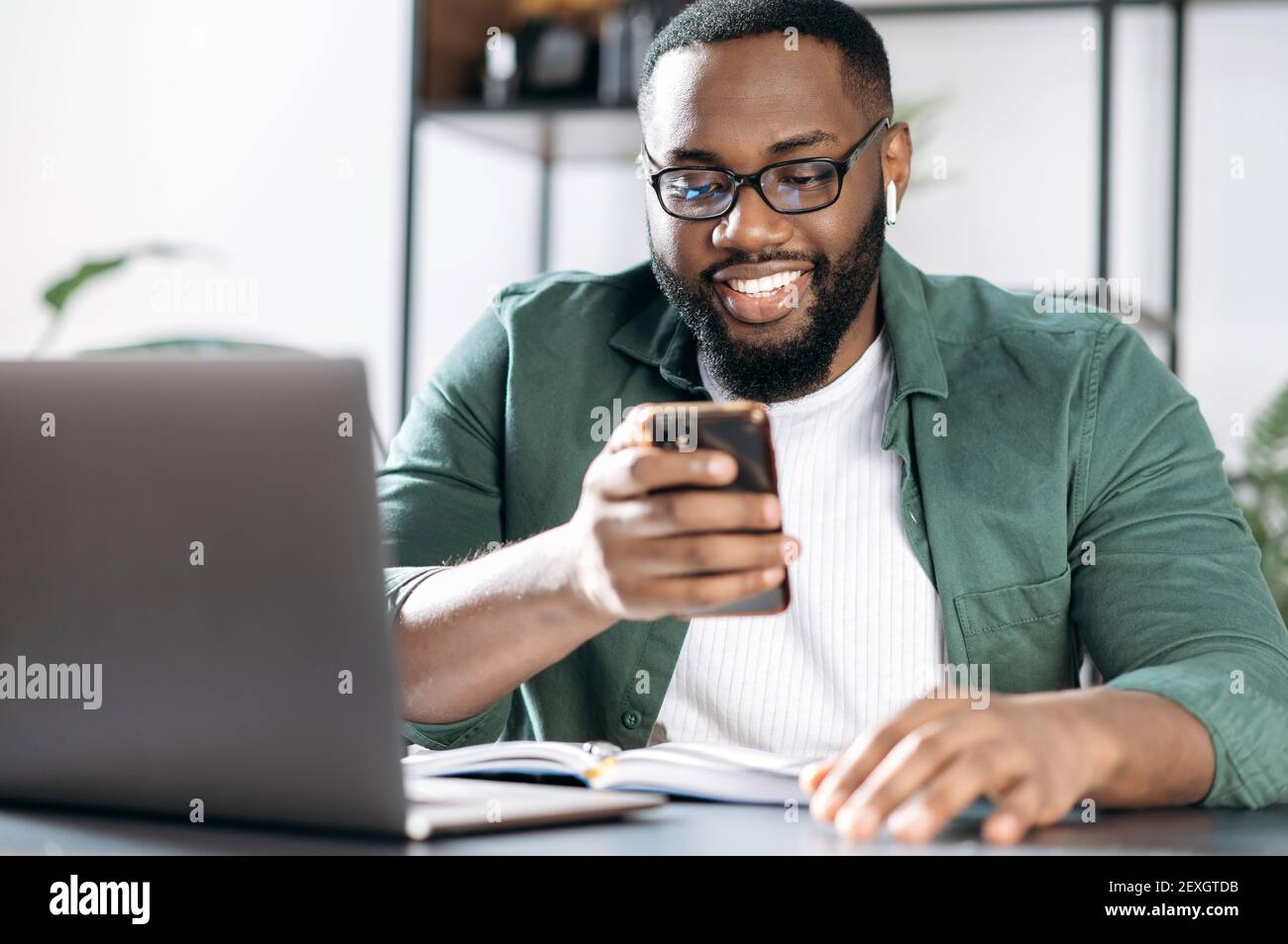 Chat online. Gioioso afroamericano ragazzo, si siede alla sua scrivania, utilizzando un telefono cellulare mentre lavora, navigare in internet, rispondere a una e-mail, chattare con un amico o la famiglia Foto Stock