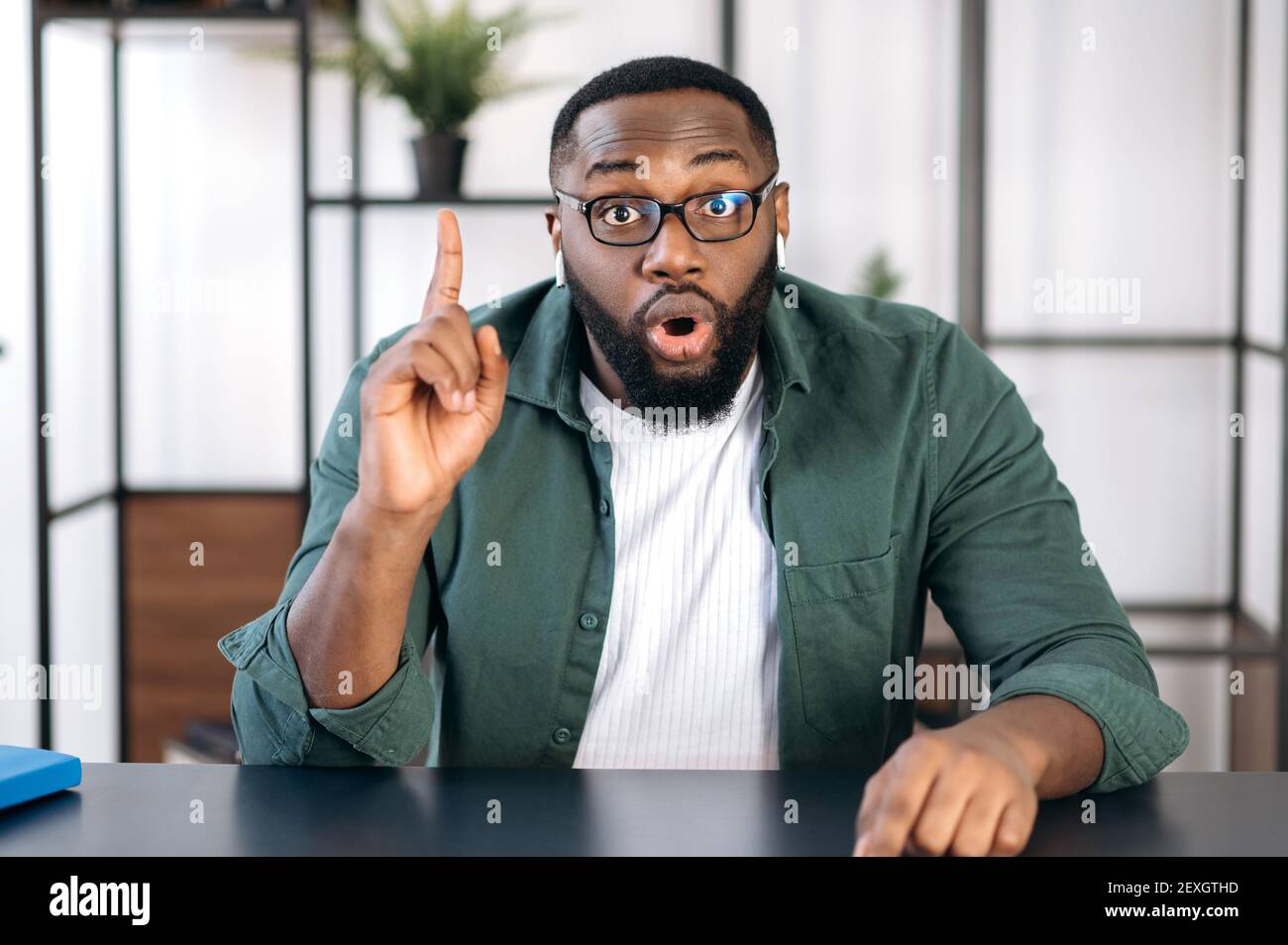 Ritratto di un ragazzo afro-americano eccitato scosso con occhiali, seduto ad un tavolo e mostrando un gesto di idea, guardando direttamente nella fotocamera Foto Stock
