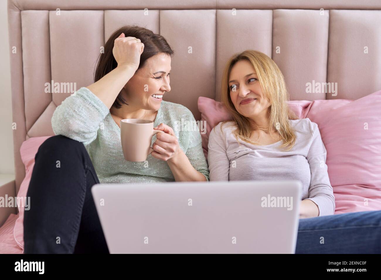Due donne mature che guardano un notebook seduto a casa letto Foto Stock