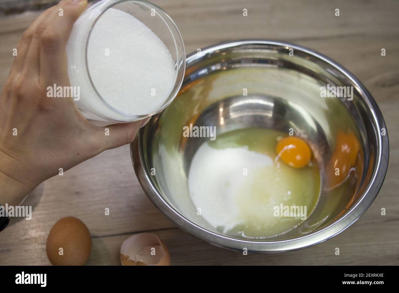 La mano aggiunge zucchero al recipiente metallico con uova e zucchero. Il secondo passaggio per preparare muffin, torta, biscotto. Cottura graduale. Aggiunta di zucchero, ad esempio Foto Stock