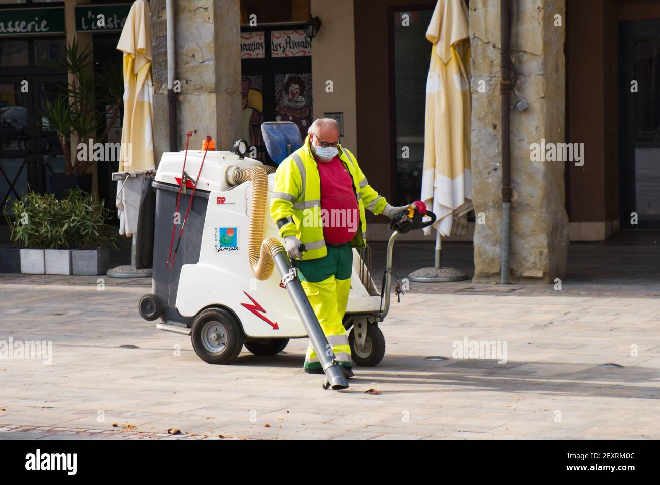 Limoux Aude France 03.04.21 uomo che indossa maschera facciale e un lavoro ad alta visibilità che riflette le revisioni tirando una macchina per la pulizia stradale sottovuoto. Centro città lastricato. Foto Stock
