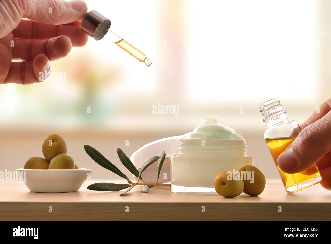 Mani con pipetta con siero e crema con estratto di oliva per la pelle con rametto e olive su tavola di legno. Vista frontale. Foto Stock