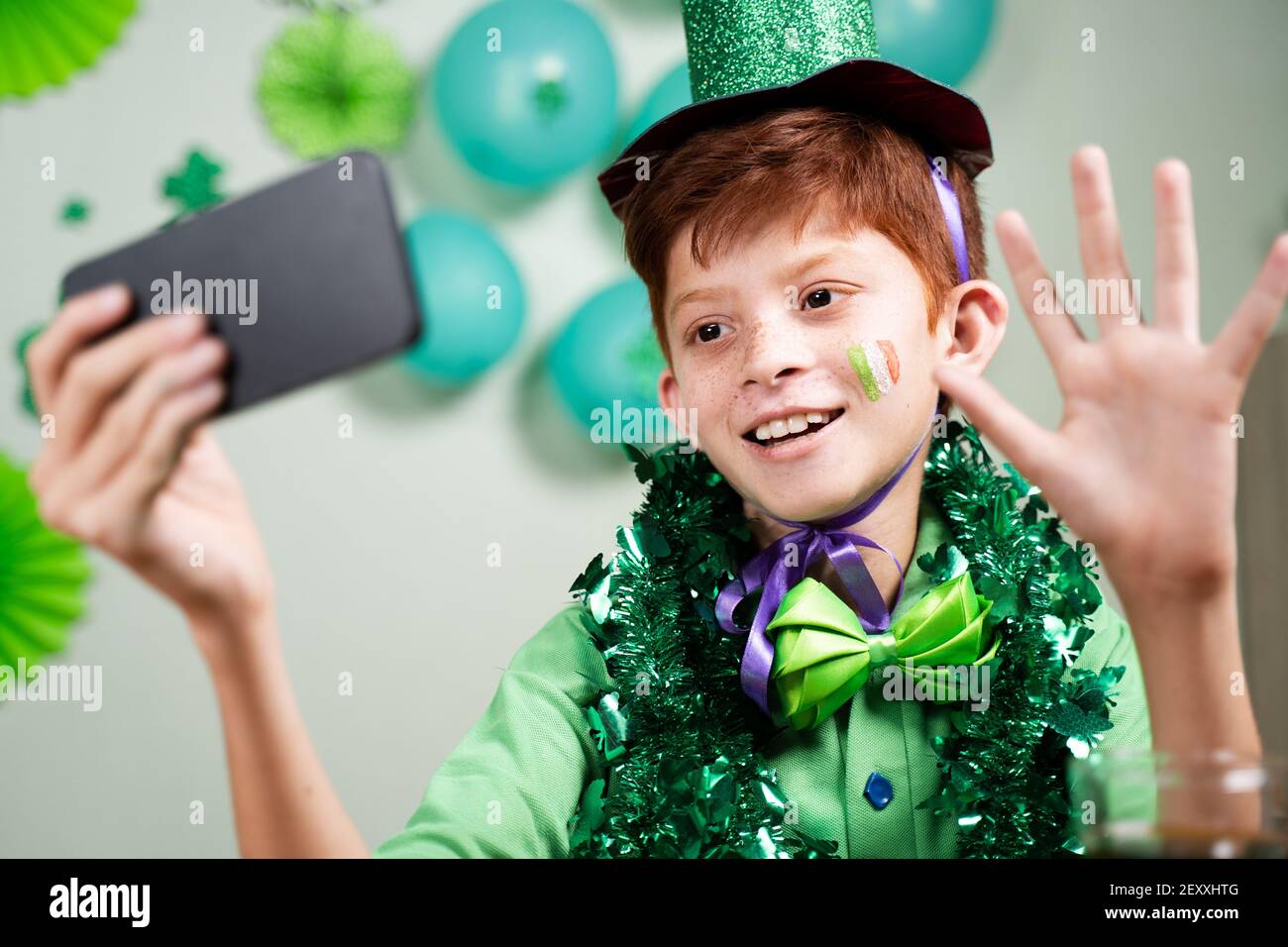 Bambino giovane in abbigliamento verde che celebra san patricks giorno dopo effettuare una videochiamata sul telefono cellulare su sfondo decorato durante coronavirus covid-19 Foto Stock