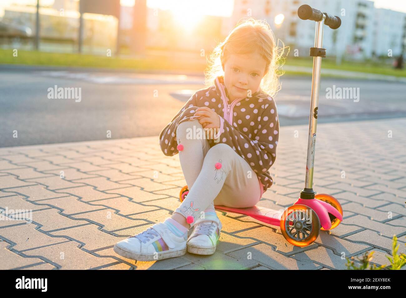 Ritratto esterno di cute bionda capelli bambina sedersi sopra kick scooter su una strada con soleggiato sfondo sfocato Foto Stock